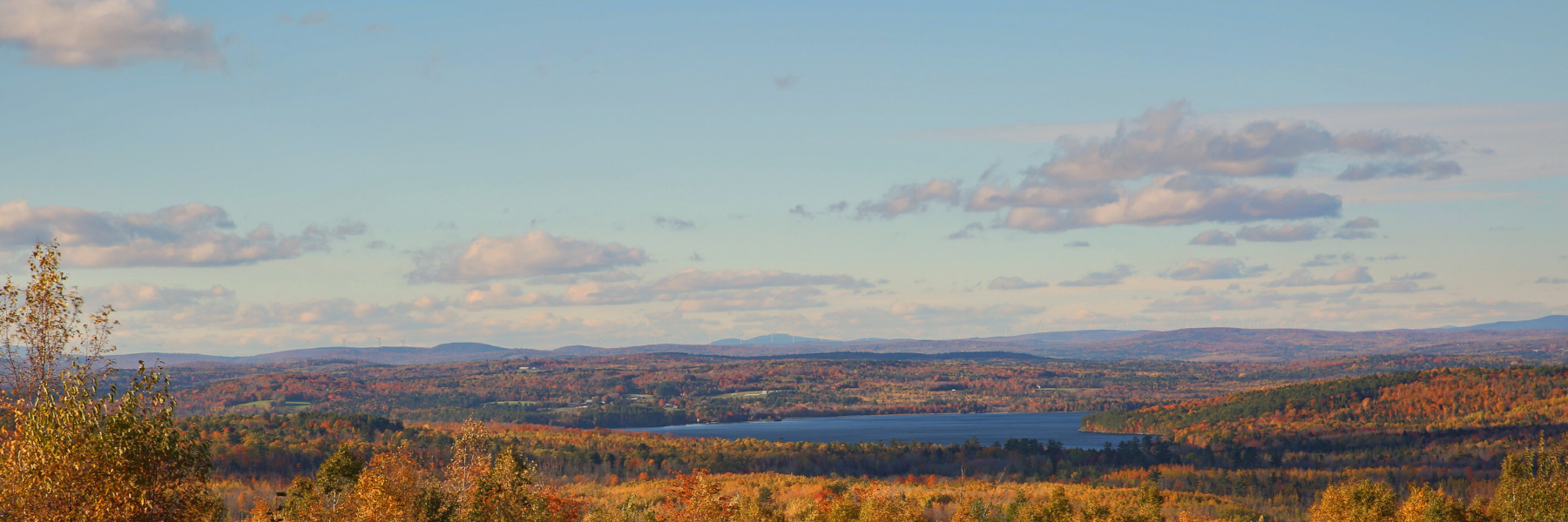 Fall in Maine landscape photograph of blue sky, lake, and trees with fall leaves. There are mountains on the horizon in the distance.